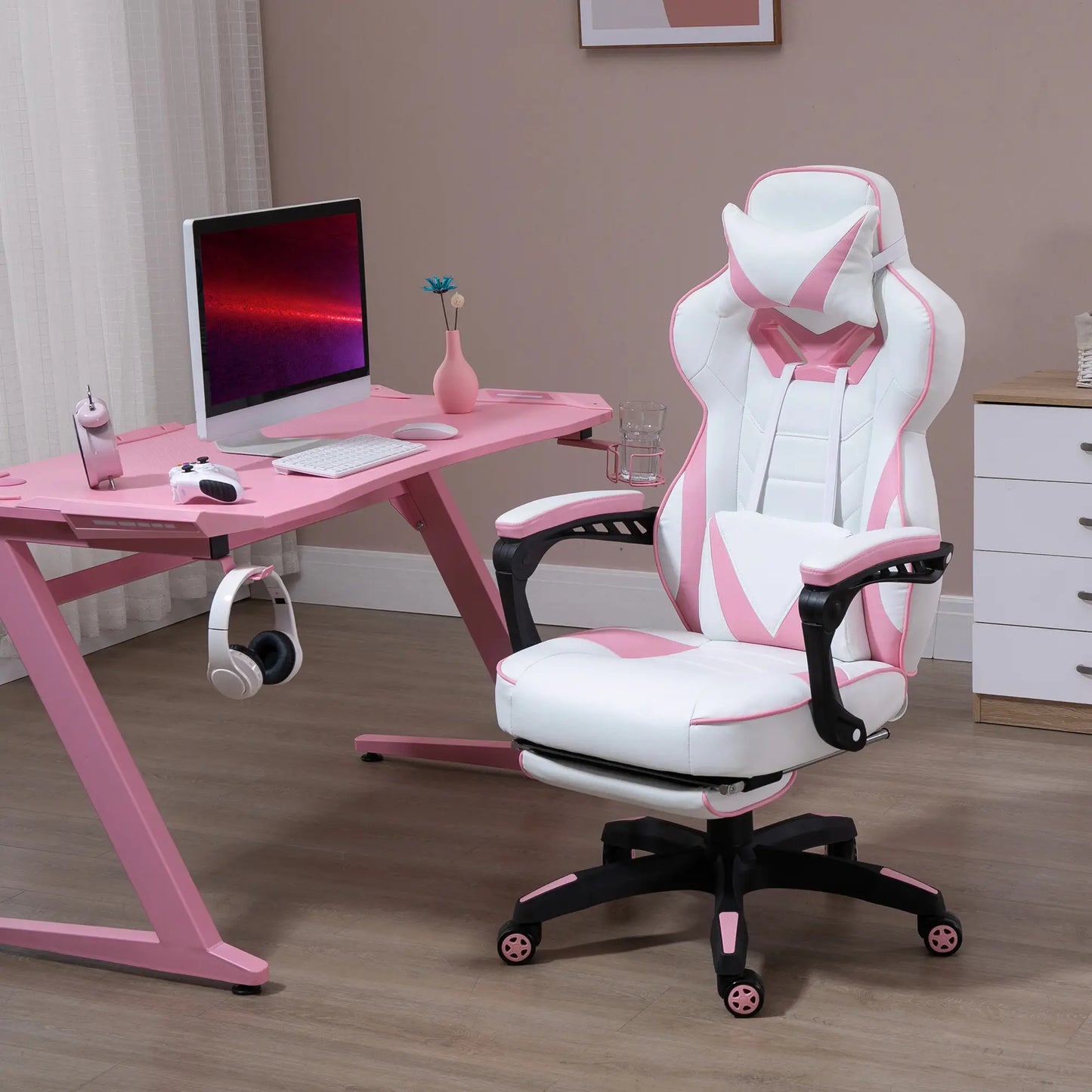 A pink and white computer gaming chair with lumbar support and footrest, positioned in a room with a pink desk and a computer setup.