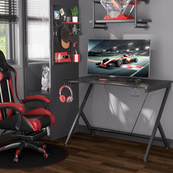 A black gaming desk with a computer setup on it, placed in a room with a racing chair, shelves of books and gaming accessories, and a window.