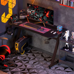 A black gaming desk with a steel frame, featuring a large worktop, a cup holder, and a cable organiser. The desk is in a room with gaming equipment setup.