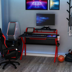 A gaming desk with a black tabletop and red metal frame, equipped with a monitor stand, wire basket, and a cup holder. The desk is in a room with a gaming chair, a basketball, and various decor.