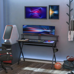 A black gaming desk with a raised monitor shelf, side hooks, and a large flat tabletop, arranged with a computer monitor, keyboard, and a gaming chair in the room.