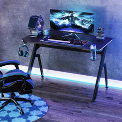 A black gaming desk with a carbon fibre surface, featuring LED light strips, a headphone hook, controller rack, and a cup holder. The desk is shown with a gaming setup, including a monitor, keyboard, and mouse.