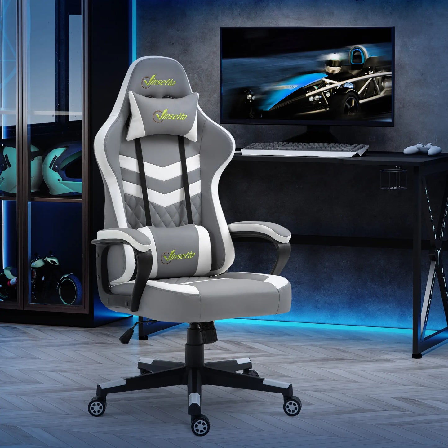 A racing style gaming chair with gray PVC leather, black fixed armrests, and extra pillows for lumbar and head/neck support. The chair is in a room with LED lights and a computer setup in the background.