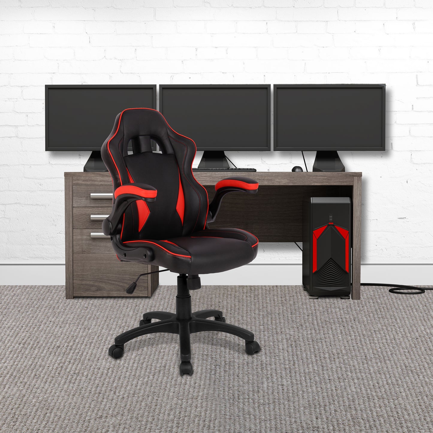 Black and red gaming chair in front of a desk with multiple monitors on a white brick wall background.