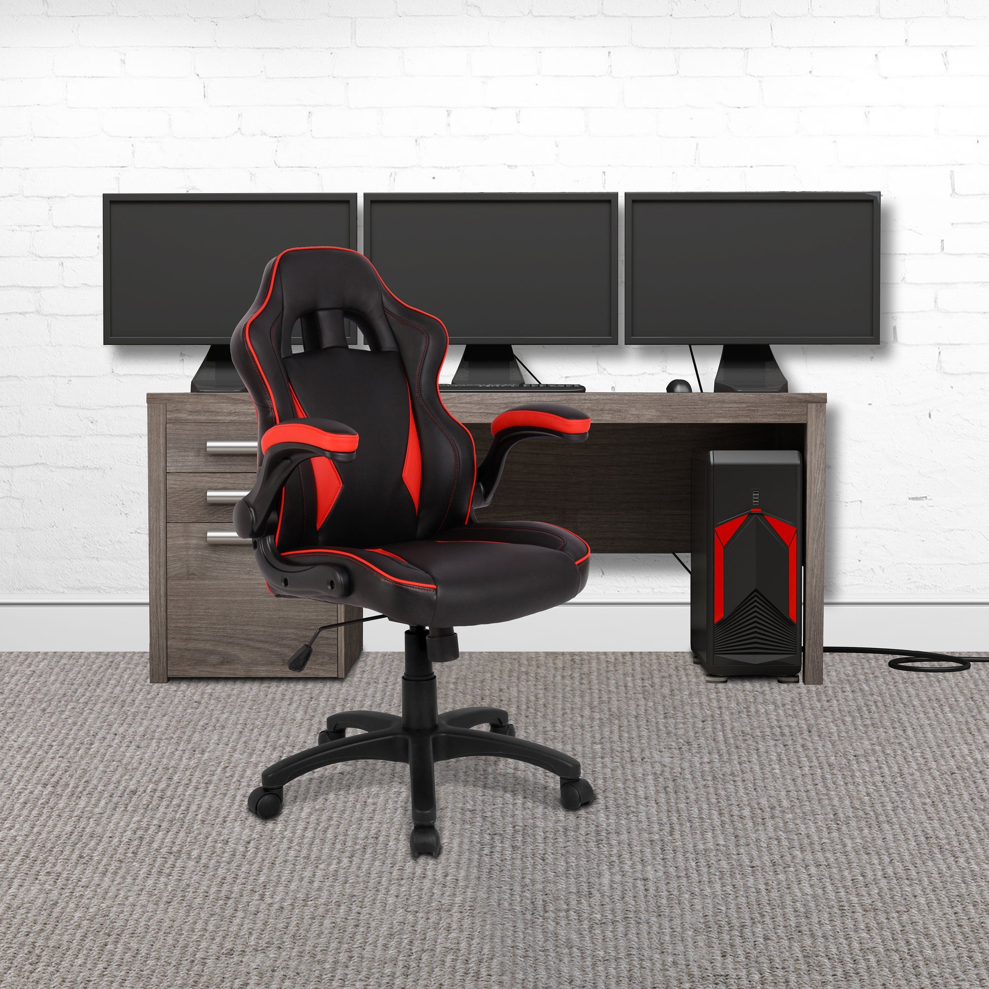 Black and red gaming chair in front of a desk with multiple monitors on a white brick wall background.